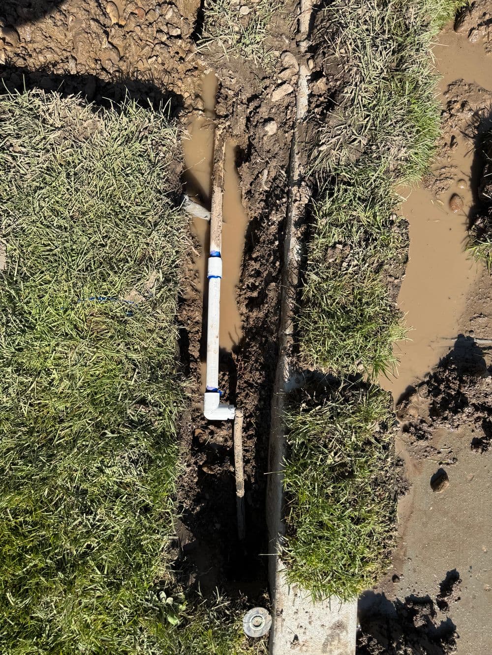 Exposed underground plumbing repair with PVC pipe in muddy trench and grassy edges.